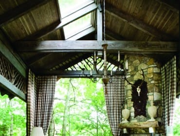 Covered porch with wood-paneled ceiling, stone fireplace, and wicker furniture. A dining set is visible on the right, and a sofa and coffee table on the left.