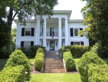White Greek Revival house with a grand portico supported by four columns, black shutters, and a manicured lawn with symmetrical hedges.