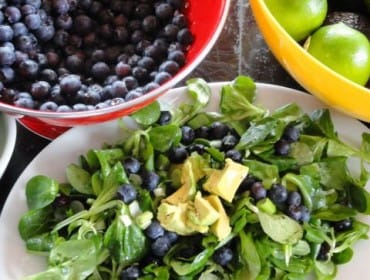 Salad with blueberries and avocado on a white plate, with a red colander of blueberries and a yellow bowl of limes and avocados in the background.