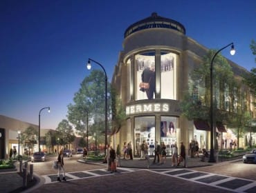 Corner view of a luxury retail building at dusk, featuring the Hermes store with illuminated signage and window displays. Pedestrians and cars are visible on the street and sidewalk.