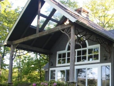 Dark gray shingled house with a covered porch featuring a skylight roof, rustic wood beams, and tree branch-like accents. Large windows and flowering bushes in the foreground.