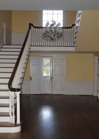 Foyer with a grand staircase, dark wood floors, yellow walls, white wainscoting, and a crystal chandelier above double doors.