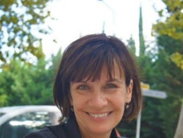 Meg Harrington smiles while sitting at a table outdoors, wearing a black collared shirt and multiple bracelets.