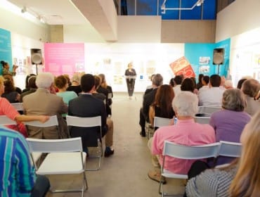 Audience seated in chairs facing a speaker at a podium in a gallery or event space with colorful display panels on the walls.