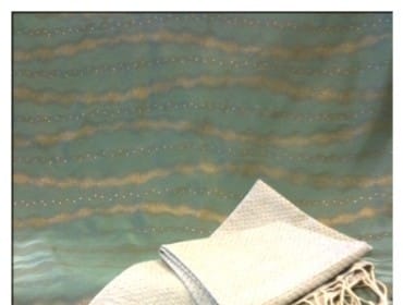 Light gray woven scarf with a textured pattern and cream-colored fringe, laid on a teal fabric with gold wavy stripes.