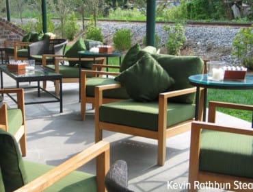 Outdoor seating area with wooden chairs and green cushions, glass-top tables, and a view of train tracks in the background.