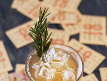 Close-up of a cocktail with ice and a sprig of rosemary, surrounded by vintage-style tickets with red numbering and text.