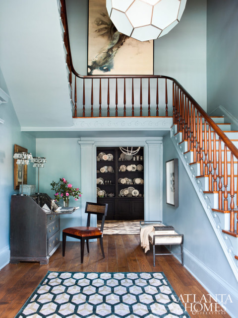 The foyer is painted in a blue-gray shade for a serene aura and punctuated by a Worlds Away honeycomb pendant; just beyond is a painted Belgian apothecary stocked with pewter.