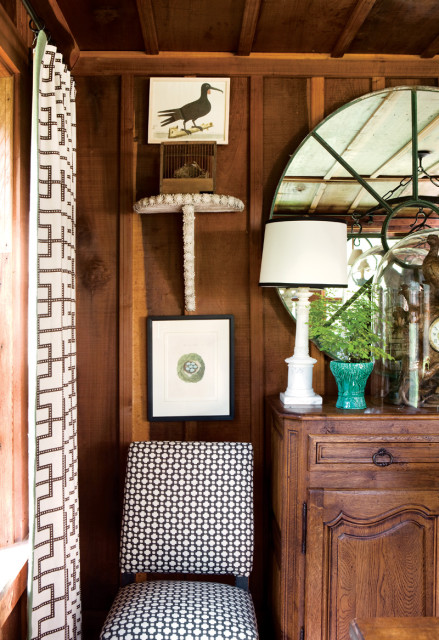 Wooden paneled room with a black and white patterned chair, botanical prints of a bird and nest, and a white lamp on an antique wooden cabinet.