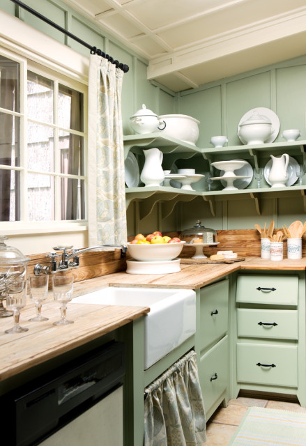 Sage green kitchen with white farmhouse sink, butcher block countertops, open shelving displaying white dishware, and a window with patterned curtains.