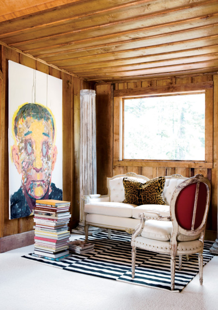 Living room with wood-paneled walls and ceiling, featuring a large abstract portrait, a white tufted sofa with a leopard print pillow, a distressed armchair with red upholstery, and a black and white striped rug.