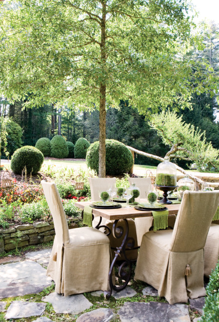 Outdoor dining table set with green-fringed placemats and moss-filled glass cloches, surrounded by manicured boxwood shrubs and lush greenery under a large tree.
