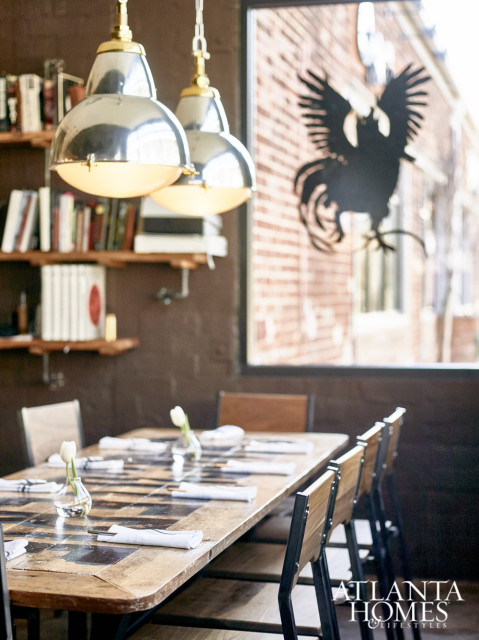 Dining table with a patchwork wood top, set with white napkins and small vases of tulips. Industrial-style pendant lights hang above, and a black rooster silhouette is visible in the window behind.