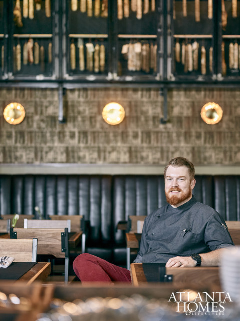 Chef with red beard wearing a grey chef's coat and red pants, sitting in a restaurant with dark leather banquettes and wooden tables.