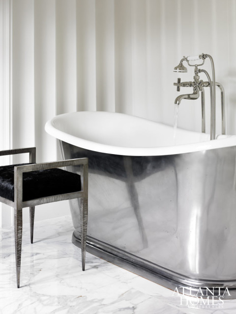 Modern bathroom with a freestanding polished metal bathtub, a black upholstered bench with a metal frame, and striped wallpaper. Water flows from a chrome faucet into the tub.
