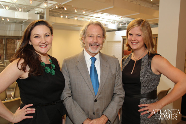 Three people smiling at the camera: a woman in a black dress with a green necklace, a man in a grey suit and blue tie, and a woman in a black and grey dress.