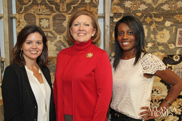 Three women smiling in front of a display of ornate rugs. The woman on the left wears a black cardigan over a white blouse. The woman in the center wears a red turtleneck sweater with a gold brooch. The woman on the right wears a cream lace top.