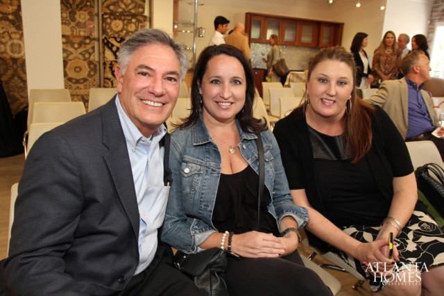 Man in a dark suit and light blue shirt smiles next to two women, one in a denim jacket and the other in a black cardigan.