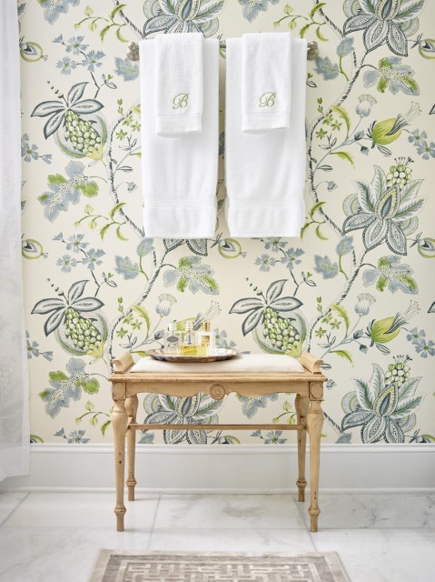 Cream wallpaper with a blue and green floral pattern, white towels embroidered with a green 'B', and a light wood bench with a cream upholstered seat.
