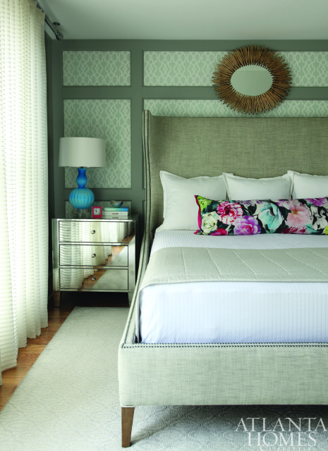 Bedroom with a tall upholstered headboard, white bedding, and a floral bolster pillow. A mirrored nightstand holds a blue lamp, and a sunburst mirror hangs on the wall.
