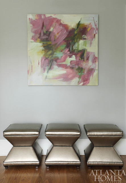 Abstract painting with pink and green hues hangs above three metallic hourglass-shaped ottomans with textured seats and dark trim.