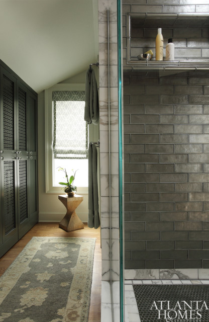 Dark green cabinetry with decorative metal inserts, a patterned Roman shade window, and a runner rug in a bathroom with a glass-enclosed shower featuring gray subway tile and marble accents.