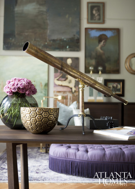 Dark wood table with brass telescope, glass vase of pink roses, gold honeycomb bowl, and tufted purple ottoman with fringe.