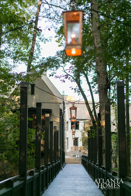 Wooden bridge with dark railings leading towards a stone building, lined with trees and hanging lanterns.