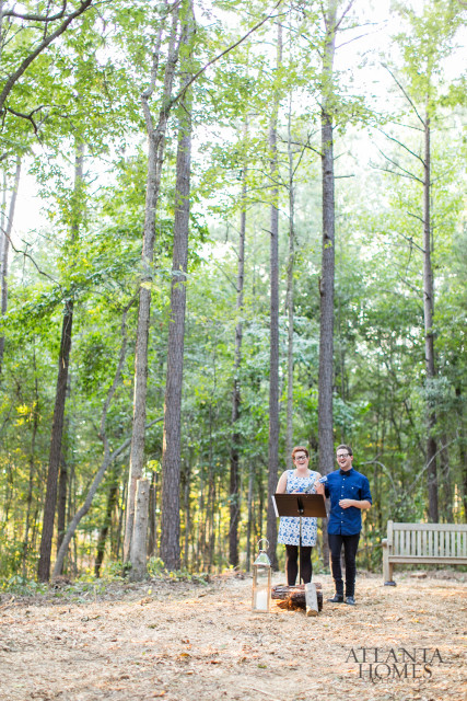 Couple standing in a wooded area with a music stand and a bench in the background. Atlanta Homes logo in the bottom right corner.