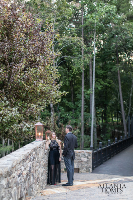 A couple stands on a stone walkway near a stone wall with lanterns, with a wooded area and bridge in the background.
