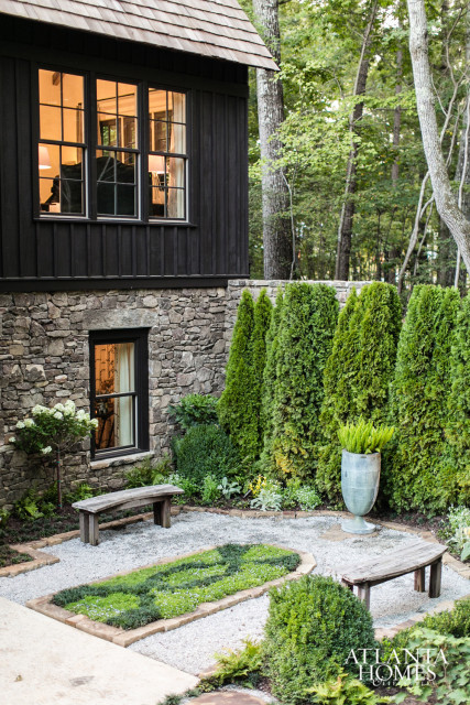 Dark wood-sided house with stone foundation, featuring a shingled roof and mullioned windows. A formal garden with gravel paths, manicured hedges, and two wooden benches sits in front.