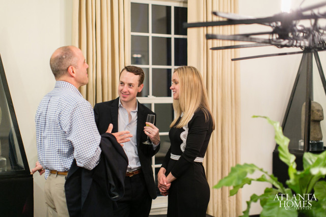 Three people in formal attire converse in a room with cream curtains, a window with white mullions, and a large abstract sculpture.