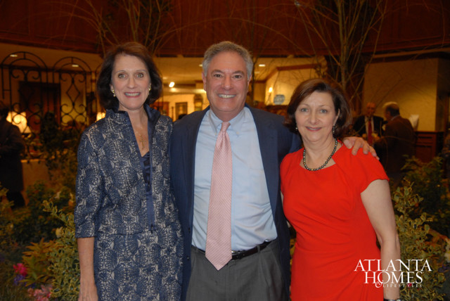 Three people posing for a photo: a woman in a blue patterned jacket and dress, a man in a blue suit jacket and pink tie, and a woman in a red dress.