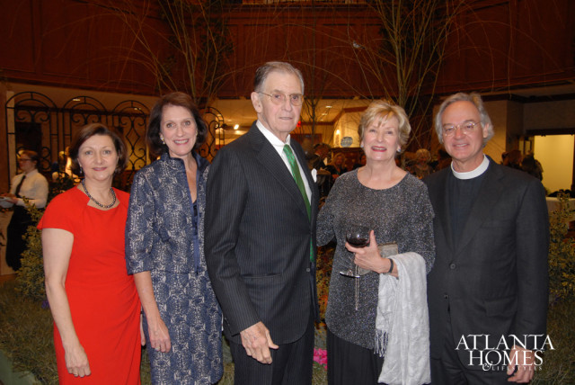 Five people pose for a photo, including a woman in a red dress, a woman in a blue patterned jacket, a man in a pinstripe suit, a woman in a sparkly top holding a wine glass, and a man in a dark suit and clerical collar.