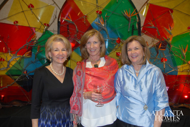 Three women smiling in front of a colorful abstract art installation with bird of paradise flowers and branches.