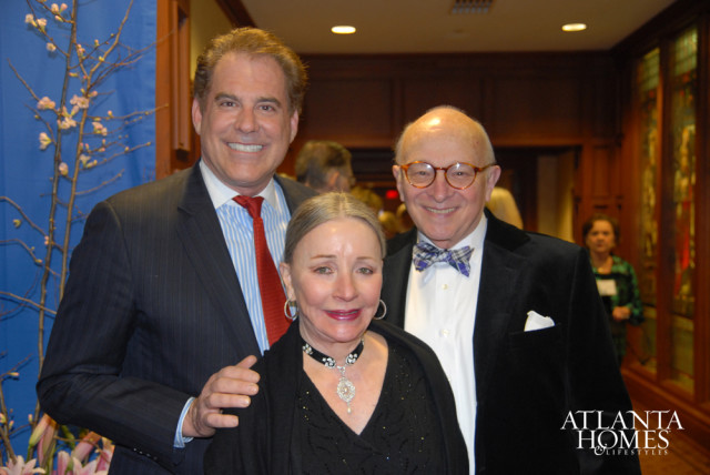 Three people posing for a photo: a man in a pinstripe suit and red tie, a woman in a black top and ornate necklace, and a man in a black velvet jacket and bow tie.