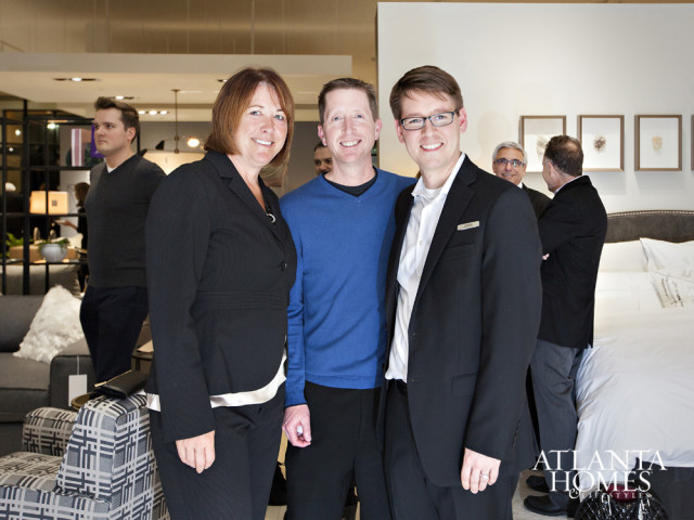 Three people smiling at the camera in a showroom with a bed and furniture in the background. A logo for Atlanta Homes & Lifestyles is visible in the lower right corner.