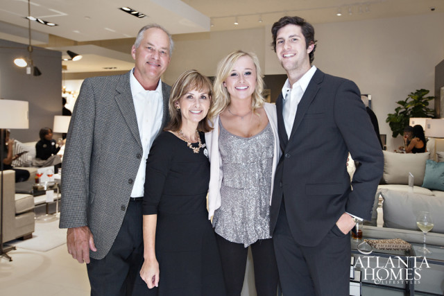 Two couples pose for a photo in a well-lit interior space with modern furniture and decor. The couple on the left consists of a man in a grey blazer and white shirt, and a woman in a black dress. The couple on the right consists of a woman in a silver sequined top and a man in a dark suit.