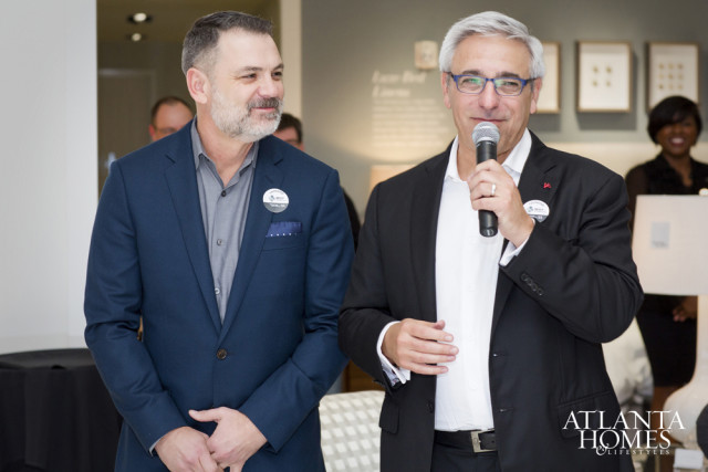 Two men in suits stand in front of a wall with framed art. The man on the right holds a microphone and speaks.