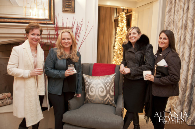 Four women in winter coats stand in a room with a fireplace, a decorated Christmas tree, and upholstered furniture.