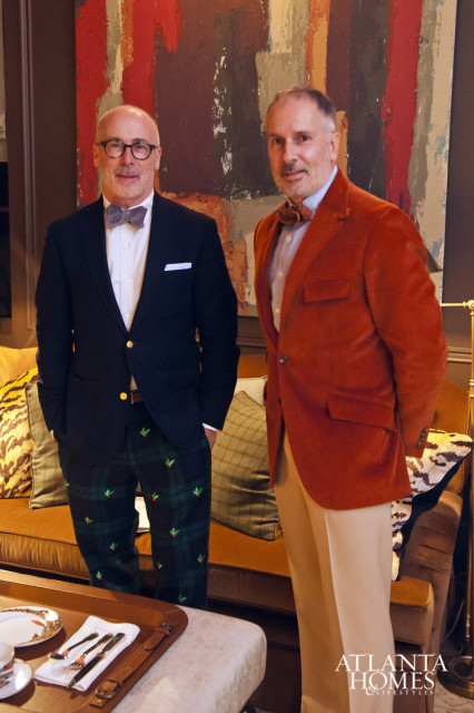 Two men standing in front of a colorful abstract painting. The man on the left wears a navy blazer, white shirt, grey bow tie, and plaid pants with holly pattern. The man on the right wears an orange corduroy blazer, light blue shirt, and tan pants.