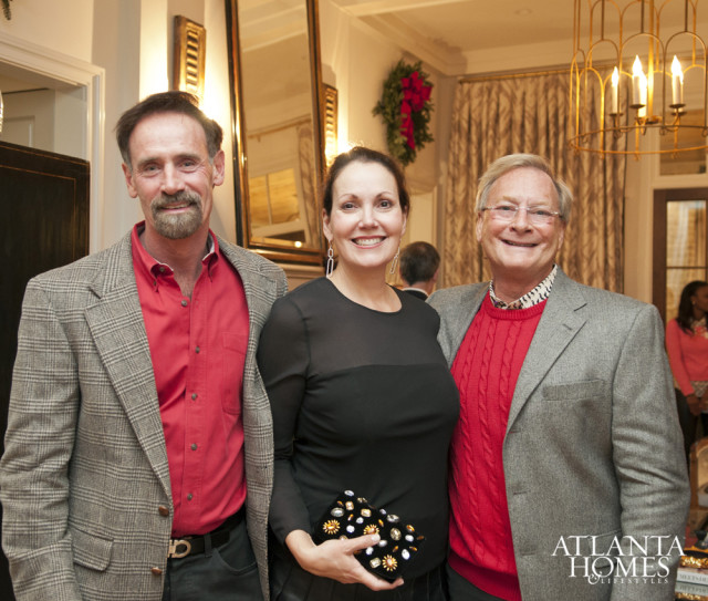 Three people posing for a photo indoors, with two men and one woman in the foreground. The woman holds a jeweled clutch.
