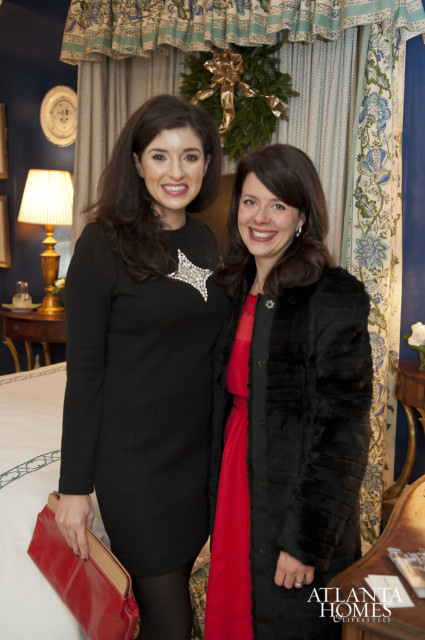 Two women pose in front of a blue wall with patterned curtains and a holiday wreath. The woman on the left wears a black dress with a star brooch and holds a red clutch. The woman on the right wears a red dress and a black fur coat.