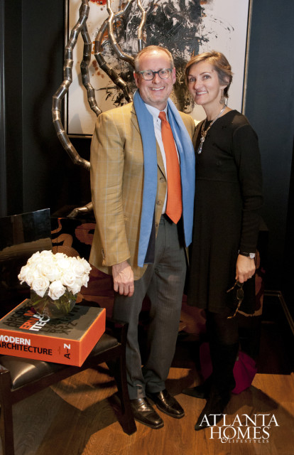 Man in tan plaid jacket with orange tie and blue scarf, and woman in black dress, pose in front of abstract art and a book titled "Modern Architecture A-Z."