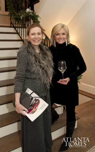 Two women smiling on a staircase decorated with holiday greenery. One woman holds a brochure with a red car on it, the other holds a wine glass.