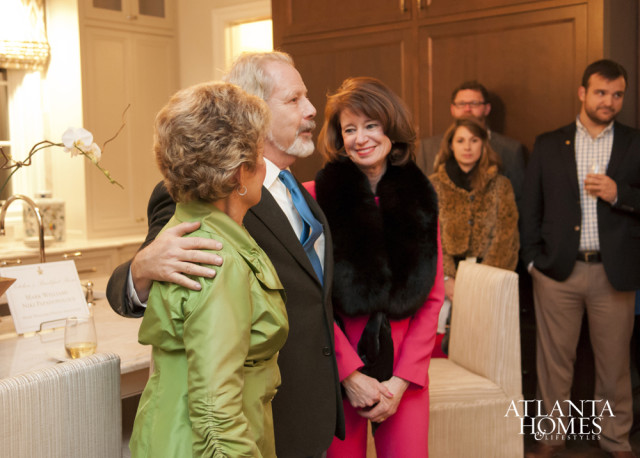 A man in a suit with a blue tie places his arm around a woman in a green jacket, while a woman in a pink suit and black fur stole smiles at him.