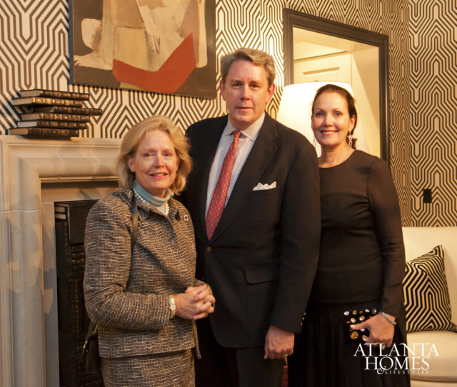 Three people stand in front of a fireplace and patterned wallpaper. The woman on the left wears a tweed suit. The man in the middle wears a dark suit and red tie. The woman on the right wears a black dress. The Atlanta Homes & Lifestyles logo is visible in the lower right corner.