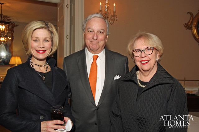 Three people posing for a photo indoors. A woman on the left wears a dark coat and pearl necklace. A man in the center wears a dark suit with an orange tie. A woman on the right wears a quilted black coat and glasses.