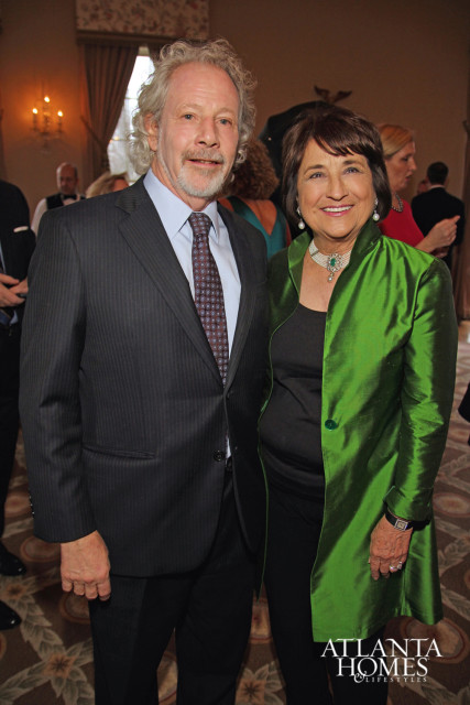 Bill Harrison and Lisa Newsom pose for a photo at an event. Harrison wears a pinstripe suit and patterned tie, while Newsom wears a green silk jacket and pearl necklace.