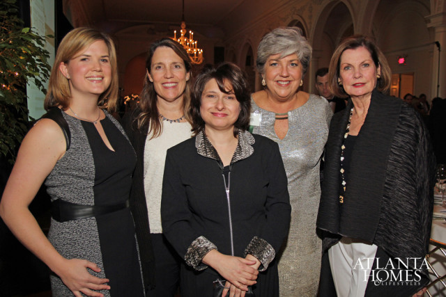 Five women pose for a photo indoors, with a chandelier and ornate architecture visible in the background. One woman on the far right wears a black shawl and a long beaded necklace.
